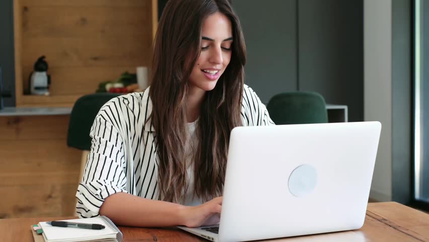 A focused young woman works diligently on her laptop, smiling while managing tasks from her comfortable home office. This productive female uses modern technology for remote job success.