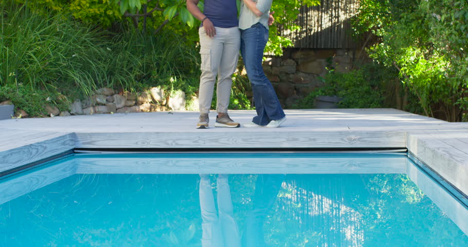 Diverse couple at pool edge, woman stepping in touching chest, man speaking to show affection. Backyard, deck, outdoor, sunlit, dappled, greenery, garden