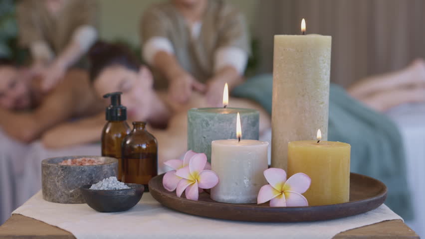 Colorful candles lit at spa center. Close up of a set of candles with aroma oil and salt at side table while couple having massage in background. Scented aroma therapy session with organic and essential oils.