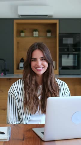 A cheerful woman engages in remote work or online learning, comfortably seated with her laptop in a contemporary home kitchen. Her bright smile conveys productivity and satisfaction.