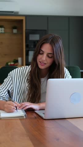 A happy young woman diligently works from her home office, smiling while taking notes and interacting with her laptop. This scene portrays remote business, online learning, and productive work.