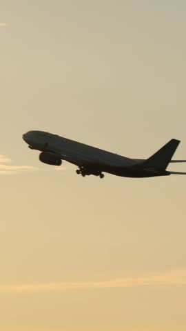 Vertical footage of airplane gaining height after taking off. Silhouette of jet flying at sunset against golden sky and bright sunlight