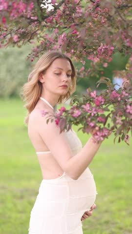 Side view of a pregnant woman in a white dress gently touching her belly beside pink blossoms in a peaceful spring garden with soft green bokeh and natural