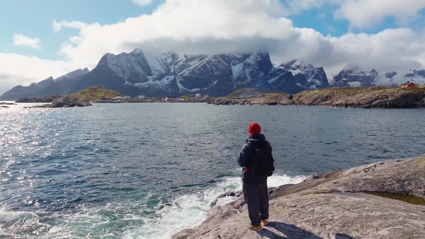 Person in red hat standing on rocky shoreline overlooking a choppy coastal bay and snow-capped