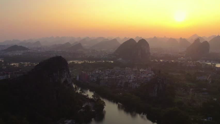 A serene dusk landscape featuring karst peaks, a winding river, and a settlement under a glowing sunset sky.