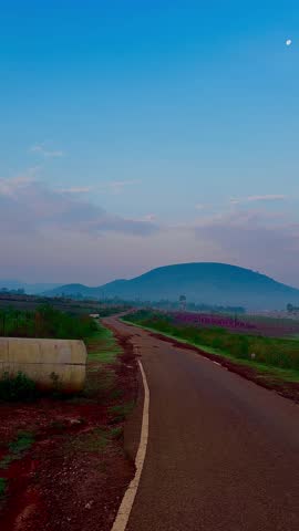 Scenic landscape of a country road passing through rural fields with foggy mountains and a calm blue sky at dawn.