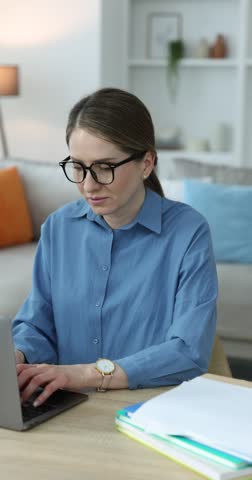 Workaholic. Overwhelmed woman in glasses at wooden desk with laptop and stack of documents indoors. Camera moving forward