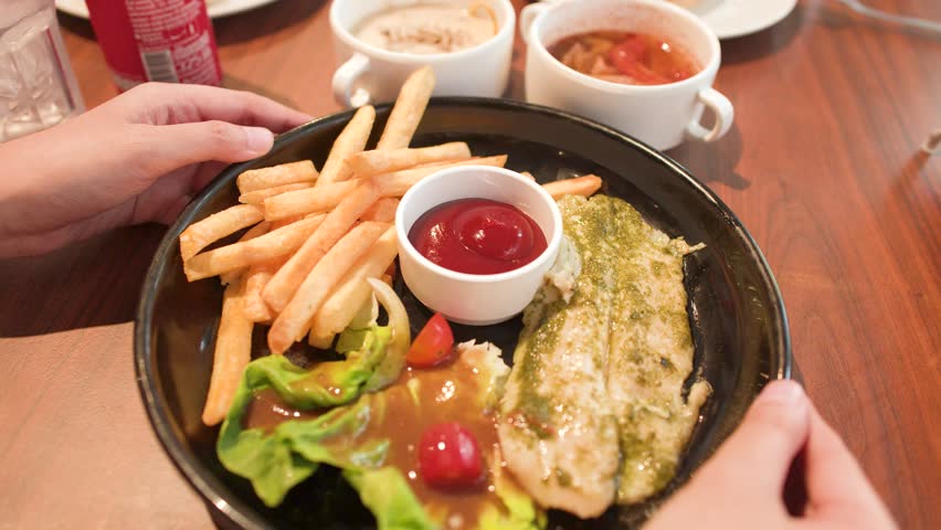 A person rotates a black plate containing grilled fish, french fries, and dipping sauces.