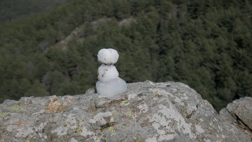 Small snowman melts on rocky ledge overlooking dense forested mountain valley