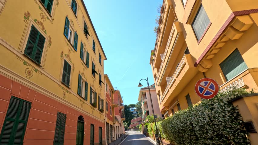 Low angle tracking shot moving through a narrow street with pastel buildings and shuttered windows.
