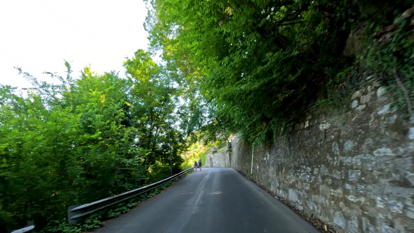 First-person perspective driving along a narrow asphalt road bordered by historic stone walls and greenery.