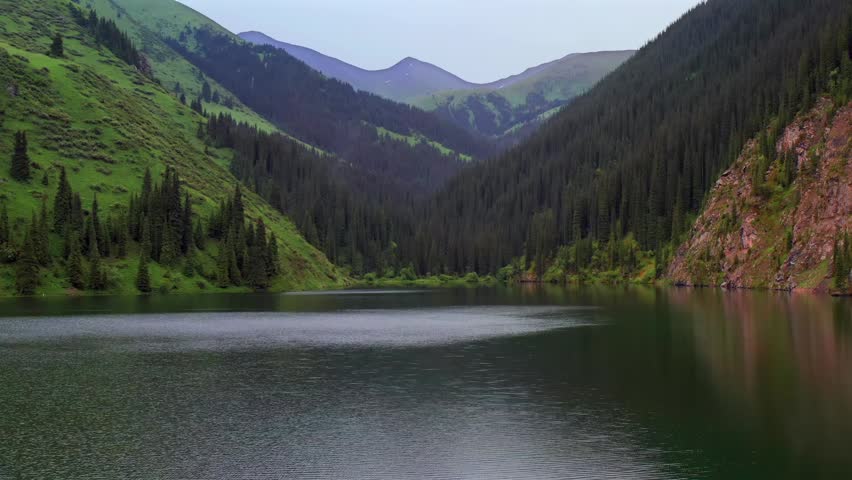Drone flying right over mountain lake, lateral movement across Kolsai lakes in Kazakhstan with green slopes and rocky cliffs, calm water surface forming travel scenery and remote nature landscape