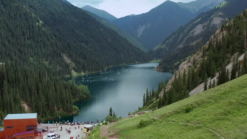 Drone flying forward over mountain lake, smooth motion across Kolsai lakes in Kazakhstan with forested slopes and steep ridges, calm water forming travel scenery and remote nature landscape