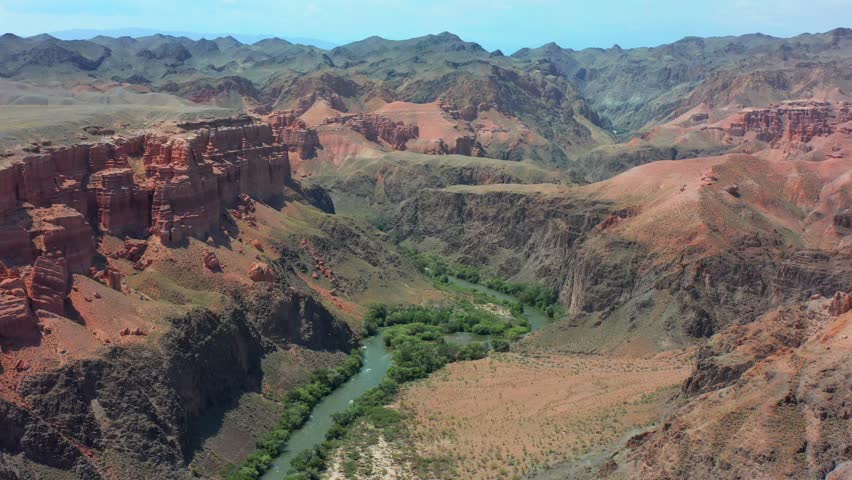 Drone flying forward above Charyn Canyon Kazakhstan, revealing winding river through wide valley, layered red rock formations and expansive mountainous terrain, dramatic natural landscape and depth