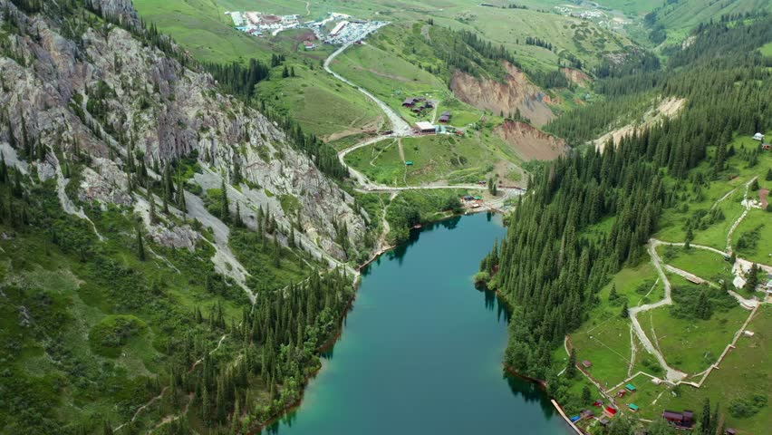 Drone flying forward over lake valley road, smooth motion across Kolsai lakes in Kazakhstan with rocky cliffs, green hills and forest, revealing travel destination and scenic mountain landscape