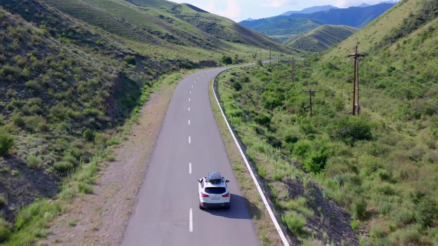 Drone tracking shot follows car driving along winding road near Zhalanash village toward Saty, surrounded by green hills and Tien Shan foothills in southeastern Kazakhstan landscape