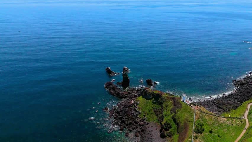 Stunning high-angle aerial drone view orbiting the iconic Seondol rock stack and a white lighthouse on the volcanic cliff of Seopjikoji, Jeju Island, South Korea.
