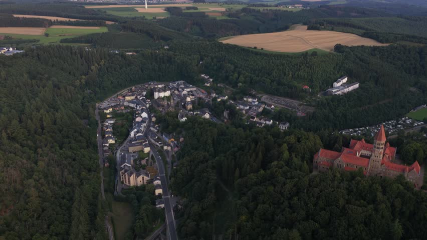 Magnificent aerial footage showing the picturesque town of clervaux and the historic saint maurice abbey, both nestled among the lush green hills and dense forests of the luxembourg ardennes
