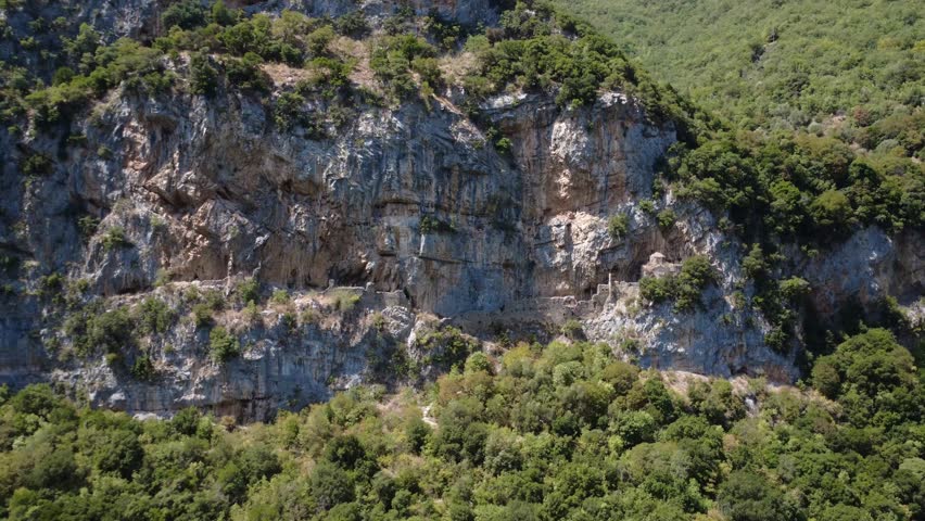 Ancient stone walls appear inside a mountain cavity surrounded by rock caves. The site lies in the Lousios River gorge in mountainous Greece with dense vegetation and large cliffs in Greece, Europe.