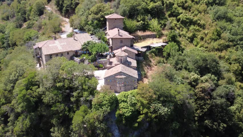 The historic and isolated Orthodox Christian Monastery of the Philosopher appears on the edge of a massive cliff inside a gorge surrounded by dense Mediterranean forest in Greece, Europe.