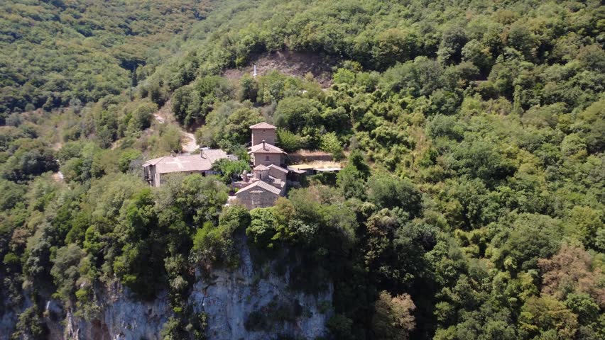 The historic Orthodox monastery rises at the edge of a massive cliff within dense forest. Stone buildings and a visible cross mark the site in Greece, Europe.