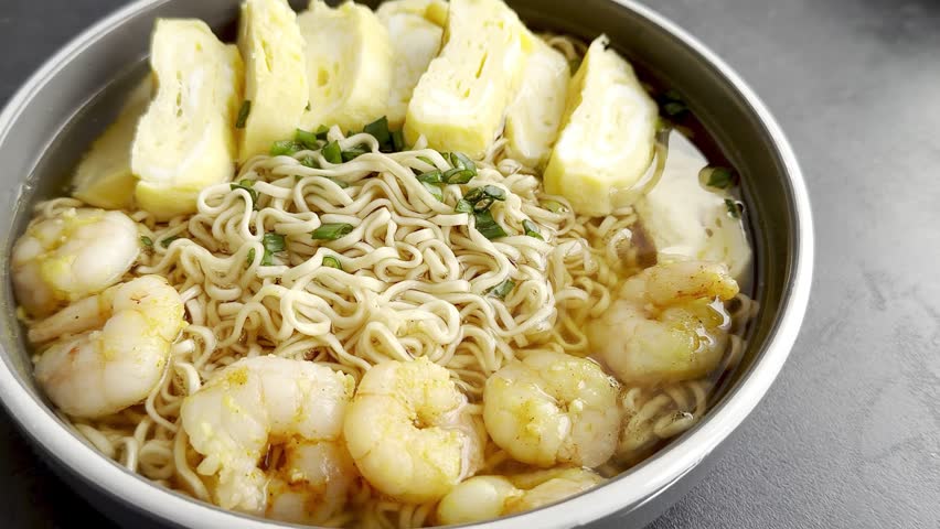 Shrimp and egg noodles in a bowl with broth, featuring cooked shrimp, sliced omelet, and green onions, presented on a dark surface