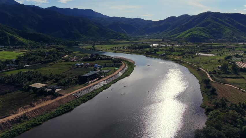 Stunning aerial drone shot of a winding river reflecting bright sunlight, set within a lush, green mountain valley. Features agricultural land and rural structures, perfect for travel and nature projects.