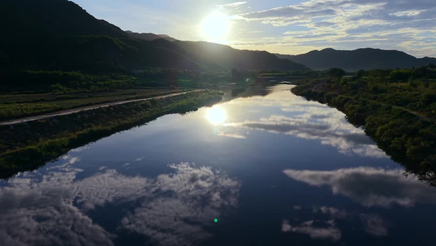 Stunning aerial drone footage of a calm river in rural Vietnam, perfectly reflecting the sky and clouds at sunset. The soft golden light highlights the peaceful mountain valley, creating a serene and captivating natural atmosphere.