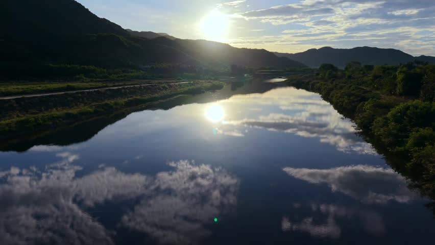 Breathtaking aerial drone footage gliding over a calm river in rural Vietnam during sunrise. The clear water perfectly mirrors the sky and clouds, while the golden morning light illuminates the surrounding lush green mountains, creating a tranquil and atmospheric landscape for travel and nature projects.