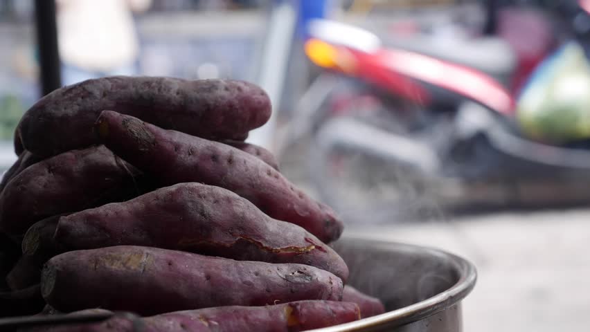 Sweet potatoes steam inside a metal container at a local outdoor market near a busy street. Traditional cooking.