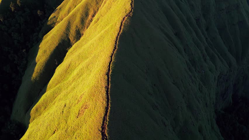 Sunlit mountain ridge trail leading toward summit across steep grassy hills in early morning light on Lombok Indonesia, tilt up aerial drone footage