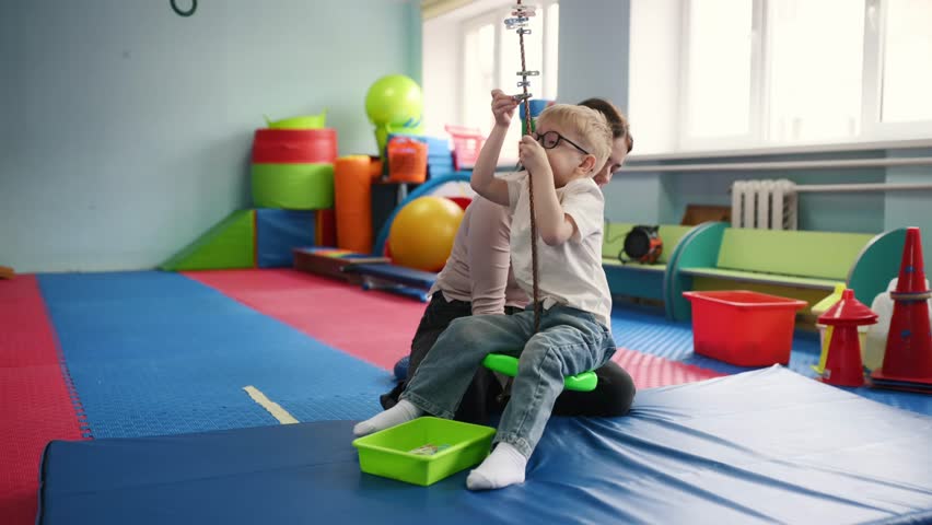 Child with glasses sitting on a swing held by a caregiver in a colorful playroom with mats, toys, and equipment visible in the background