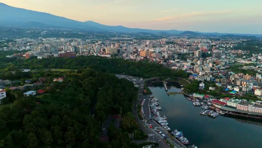 Panoramic high-angle aerial drone view orbiting right over Seogwipo Port and cityscape, revealing a lush green forest and majestic Mount Hallasan in Jeju Island, South Korea.
