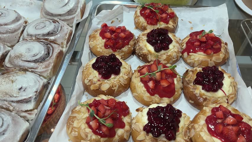 Freshly baked pastries displayed in bakery showcase. Assortment includes cinnamon rolls, homemade buns with poppy seeds, sweet pastries and traditional cottage cheese topped with berries and fruit