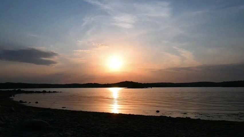 Sun is setting over the water at a beach in Norway. The sky is bright with orange and blue colors. There are distant hills and a smooth shoreline. People are enjoying the evening.