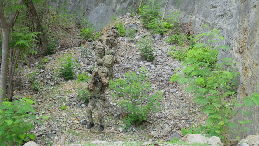 Group of armed soldiers in camouflage uniform moving through rocky canyon during military tactical operation outdoor. Special forces operators holding rifle weapons near stone cliff