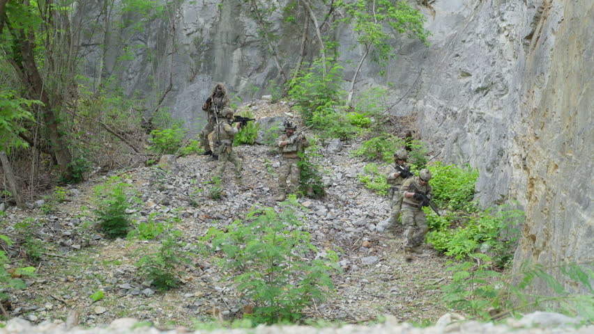 Group of armed soldiers in camouflage uniform moving through rocky canyon during military tactical operation outdoor. Special forces operators holding rifle weapons near stone cliff