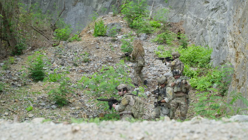 Group of armed soldiers in camouflage uniform moving through rocky canyon during military tactical operation outdoor. Special forces operators holding rifle weapons near stone cliff