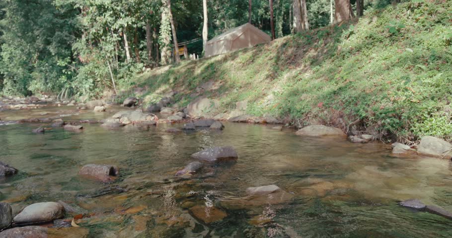 Camping tent stands on grassy riverbank next to shallow rocky stream. Sunlight filters through dense forest trees during summer outdoor adventure. Wilderness campsite in nature.