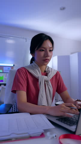 Asian woman in office using laptop with document pen on desk wearing pink shirt and sweater, focused employee typing and writing paper in modern evening workspace Vertical