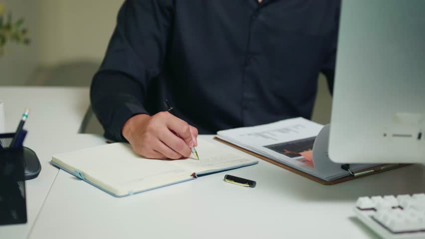 Professional man in dark shirt writing in notebook at white office desk with monitor, clipboard, and documents, ideal for business and productivity concepts.