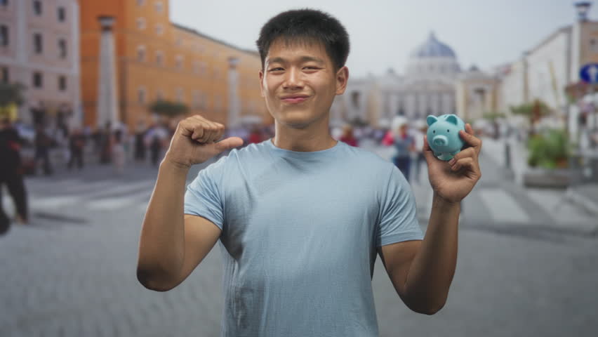 Man young wearing light blue tshirt holding small blue piggy bank and pointing thumb to chest in busy street with blurred crowd and plaza behind; saving confidence.
