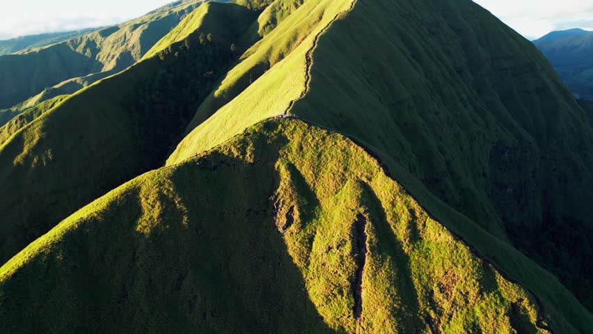 People standing on a steep mountain trail above sunlit volcanic hills and layered green mountain landscape, pullback tilt up aerial drone footage