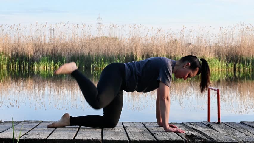 Athletic woman lifts leg doing fire hydrant exercise on wooden pier. Fit lady extends hips near calm lake for physical improvement. Active female person trains body during morning workout.