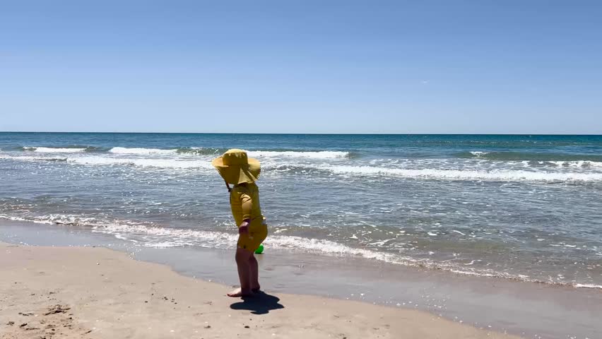 Young child in a yellow swimsuit and hat playing with a green ball on the seashore in Tarragona