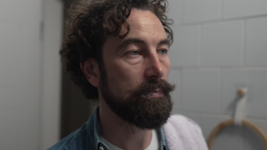 Handsome caucasian man with a beard and curly hair brushing his teeth with a toothbrush in the bathroom. Dental health, oral hygiene, and daily morning routine for a fresh clean mouth