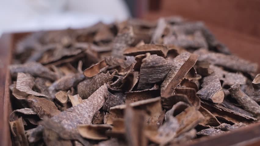 Close-up shot with shallow depth of field showing traditional dry oud wood chips resting inside a dark wooden box. The natural brown agarwood incense pieces are illuminated by soft indoor lighting.