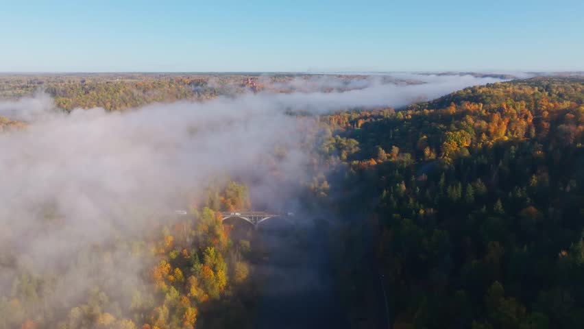 Low clouds drift across Gauja valley revealing bridge amid autumn colors