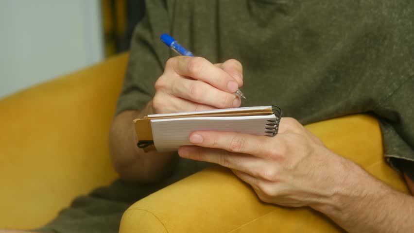 Close up man hands writing notes with blue ballpoint pen on small spiral notebook, sitting on comfortable yellow armchair, focusing on act of handwriting. Man writing with pen on small notebook.