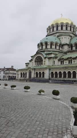 A cinematic video shot of the iconic Alexander Nevsky Cathedral, a masterpiece of Neo-Byzantine architecture. The footage captures the grand, weathered stone facade, the prominent shining golden domes, and the intricate green copper roofs against a cloudy sky. This high-quality B-roll beautifully showcases the historical and religious significance of this Bulgarian landmark, making it an excellent choice for travel documentaries, cultural heritage projects, and cinematic B-roll of Sofia.